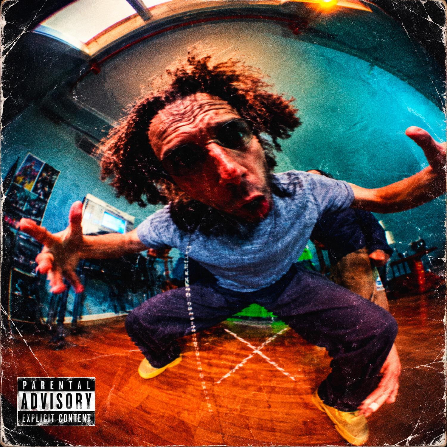 Cover art for Tell Me: a fisheye photo of a curly-haired man in a grey T-shirt crouched low on a wooden studio floor, arms flung wide and face pushed into the lens, under a curved teal wall and a skylight, a Parental Advisory label in the bottom corner